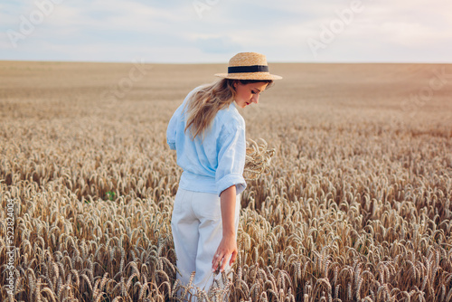 Young woman walking in summer field wearing straw hat and linen shirt holding wheat bundle. Girl touching wheat