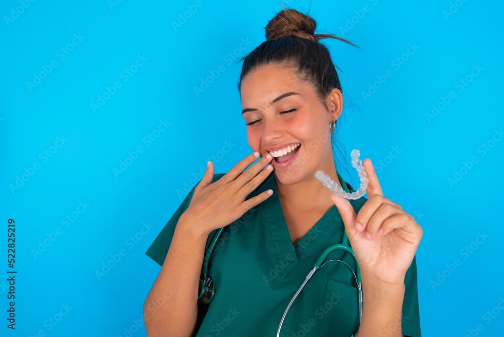 Happy beautiful doctor woman wearing medical uniform over blue background holding and showing at camera an invisible aligner while laughing. Dental healthcare and confidence concept. Happy beautiful doctor woman wearing medical uniform over blue background holding and showing at camera an invisible aligner while laughing. Dental healthcare and confidence concept.