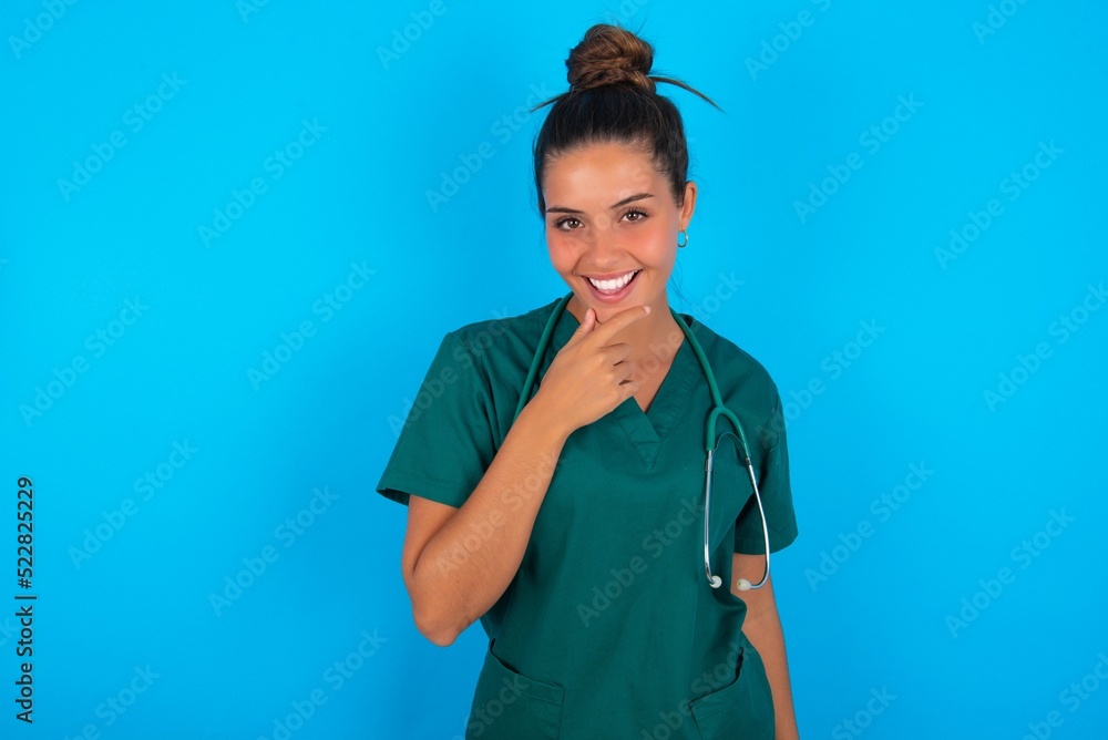 beautiful doctor woman wearing medical uniform over blue background laughs happily keeps hand on chin expresses positive emotions smiles broadly has carefree expression beautiful doctor woman wearing medical uniform over blue background laughs happily keeps hand on chin expresses positive emotions smiles broadly has carefree expression