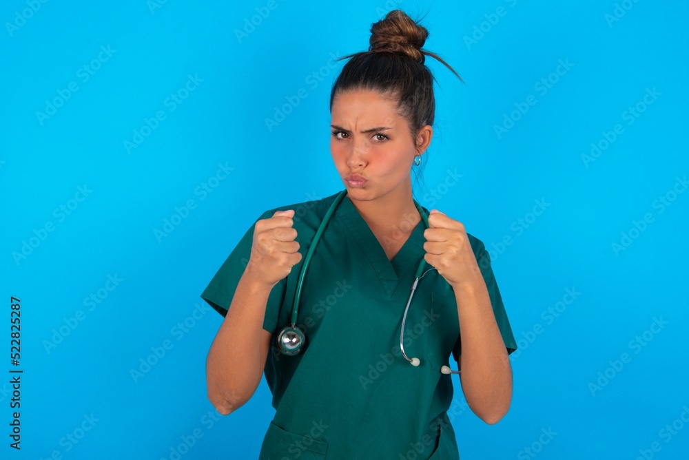 Displeased annoyed beautiful doctor woman wearing medical uniform over blue wall clenches fists, gestures pissed, ready to revenge, looks with aggression at camera stands full of hate, being pressured Displeased annoyed beautiful doctor woman wearing medical uniform over blue wall clenches fists, gestures pissed, ready to revenge, looks with aggression at camera stands full of hate, being pressured