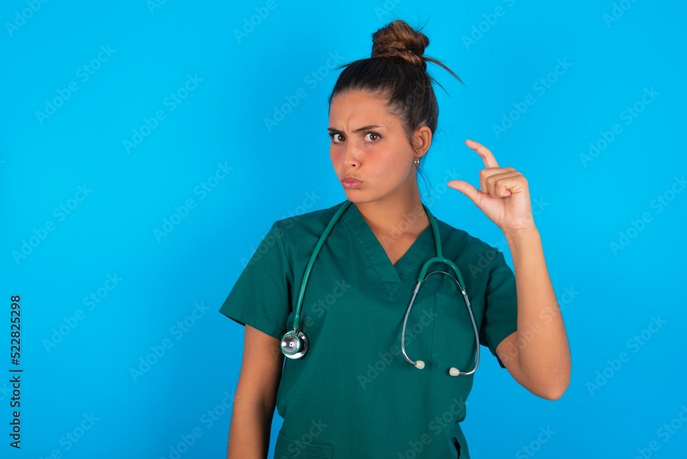 beautiful doctor woman wearing medical uniform over blue background purses lip and gestures with hand, shows something very little. beautiful doctor woman wearing medical uniform over blue background purses lip and gestures with hand, shows something very little.