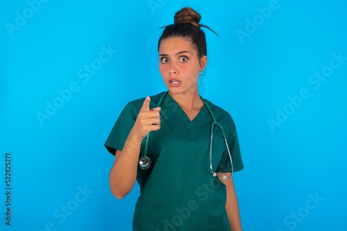 Shocked beautiful doctor woman wearing medical uniform over blue background points at you with stunned expression