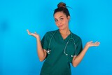 Careless attractive beautiful doctor woman wearing medical uniform over blue background shrugging shoulders, oops.