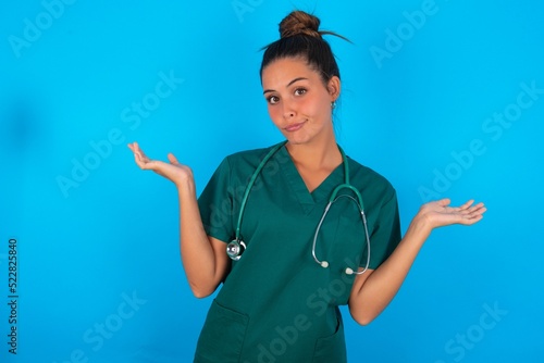 Careless attractive beautiful doctor woman wearing medical uniform over blue background shrugging shoulders, oops. photo