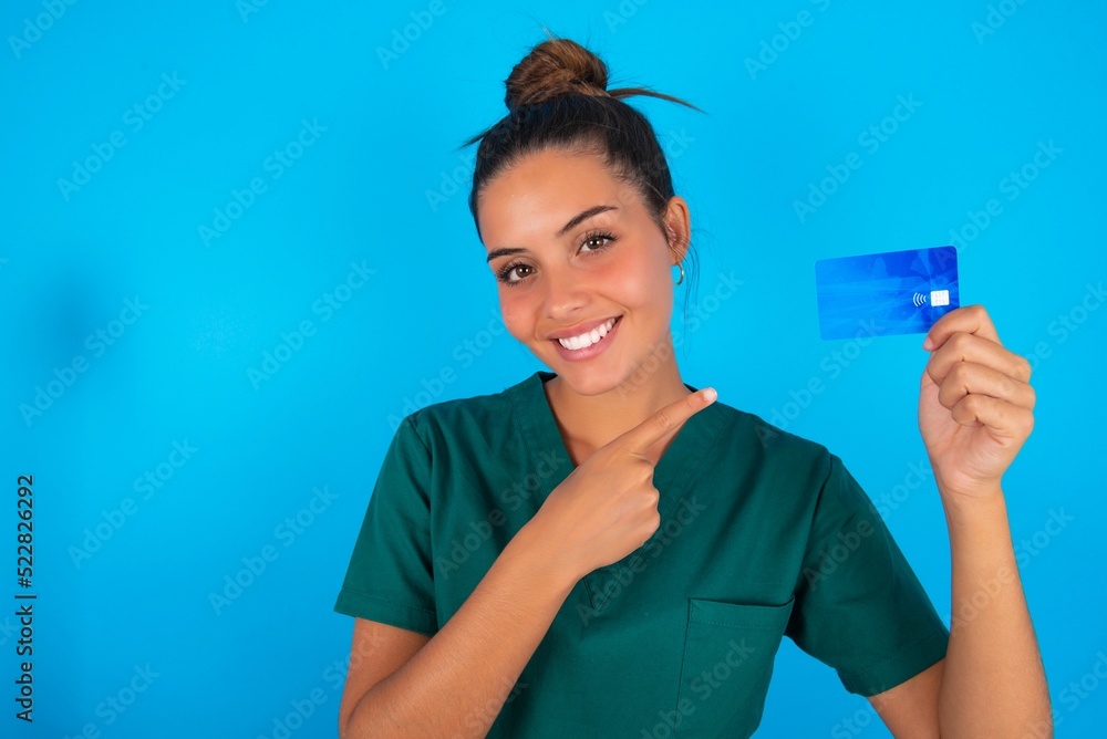 Photo portrait of beautiful doctor woman wearing medical uniform over blue background doing purchase with pointing finger credit bank card Photo portrait of beautiful doctor woman wearing medical uniform over blue background doing purchase with pointing finger credit bank card