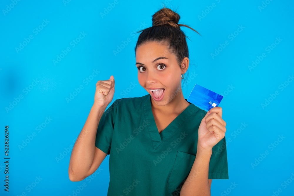 Photo of lucky impressed beautiful doctor woman wearing medical uniform over blue background arm fist holding credit card. Celebrated Photo of lucky impressed beautiful doctor woman wearing medical uniform over blue background arm fist holding credit card. Celebrated