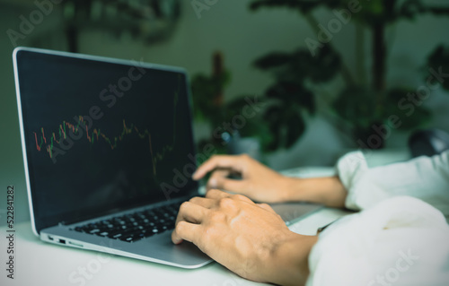 Close up, hand of businessman 30s looking stock tickers or graphs cryptocurrency trading platforms on laptop with a cup of coffee, calculator, notebook on the table at home office in the morning.