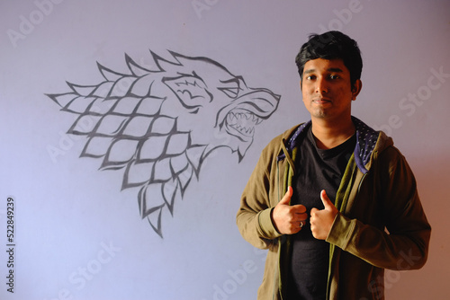 Young south asian boy wearing winter dress in front of a wall which have an art of symbol of house stark of the series game of thrones.