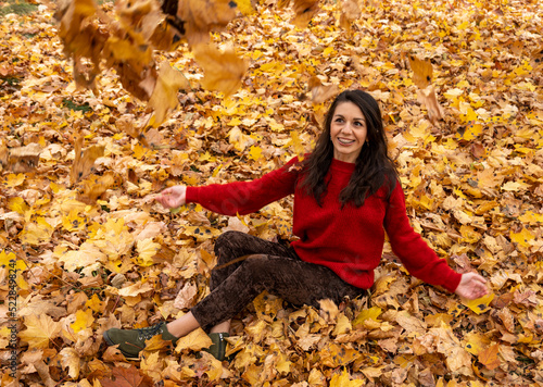 A young woman in a red sweater sits on yellow autumn foliage. A woman throws autumn leaves. Brunette woman portrait