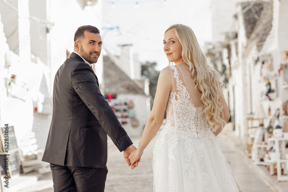 Portrait of couple walking, looking back. Happy bride wearing long white dress interlocking hands with groom in suit.