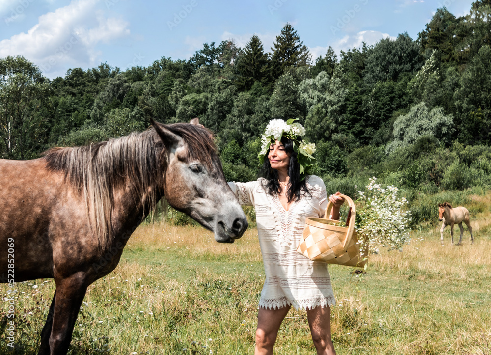 A girl in a wreath of flowers on her head and with a basket in her hands strokes a horse. The horse is grazing in the meadow. A girl in a wreath of flowers on her head and with a basket in her hands strokes a horse. The horse is grazing in the meadow.