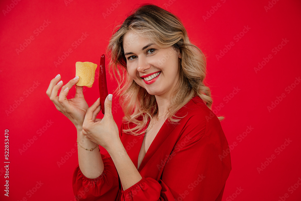Smiling pleased blonde woman showing junk chili pepper and yummy salty potato chips and look at camera on red background Smiling pleased blonde woman showing junk chili pepper and yummy salty potato chips and look at camera on red background