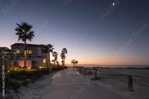 A beach path on Coronado beach, San Diego, at sunrise. The sky is turning orange and the moon can still be seen in the sky. 