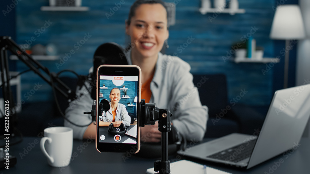 Attractive social media influencer sitting at home studio desk while starting streaming from living room with smartphone. Young content creator recording daily vlog with touchscreen modern device. Attractive social media influencer sitting at home studio desk while starting streaming from living room with smartphone. Young content creator recording daily vlog with touchscreen modern device.