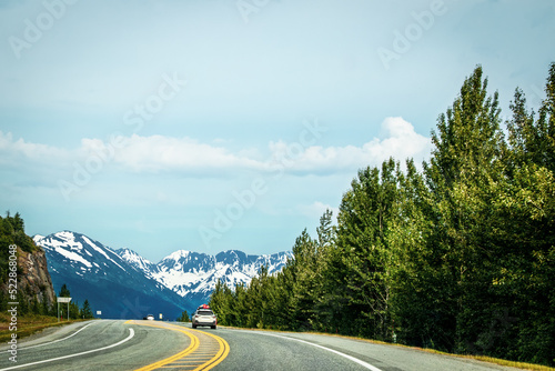 Driving from Anchorage to Seward Alaska on Highway 9 south on the Kenai Peninsula with snowcovered mountains in the distance and evergreen trees on the sides - SUV with gas tanks on top ahead on paved photo