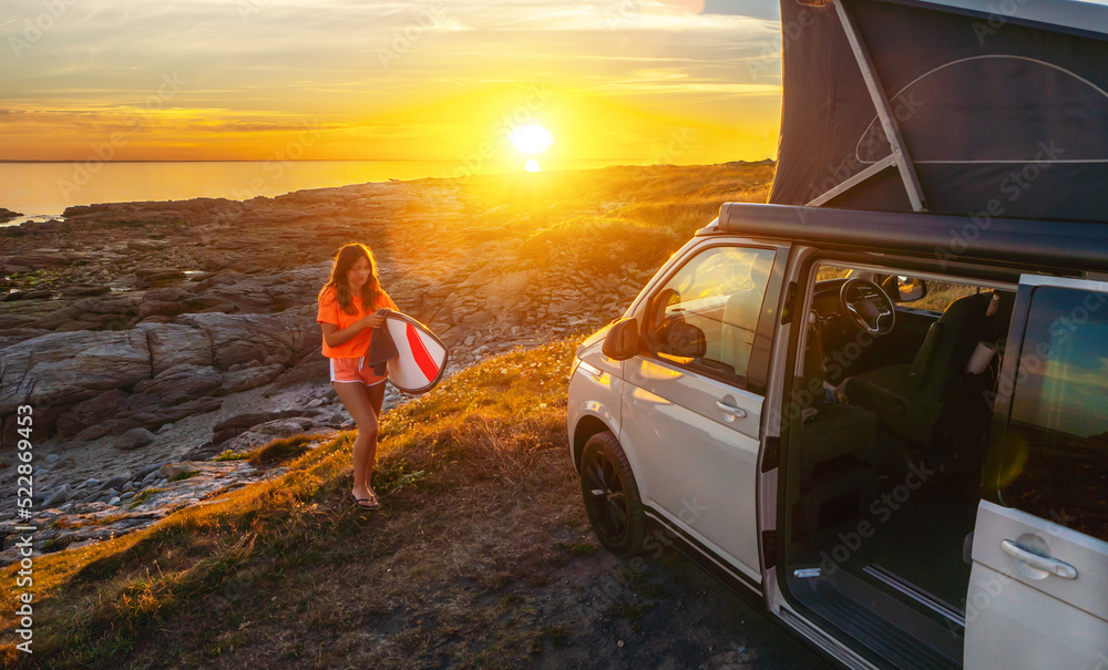 Surfer girl walking near her mini van and looking on the ocean at summer sunset with a surfboard on her side Surfer girl walking near her mini van and looking on the ocean at summer sunset with a surfboard on her side