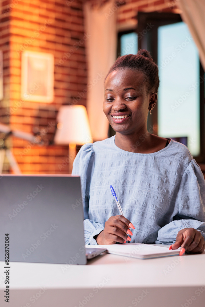 Smiling female student watching educational tutorials on laptop and taking notes in notebook. Young african american woman attending remote university online courses on internet Smiling female student watching educational tutorials on laptop and taking notes in notebook. Young african american woman attending remote university online courses on internet