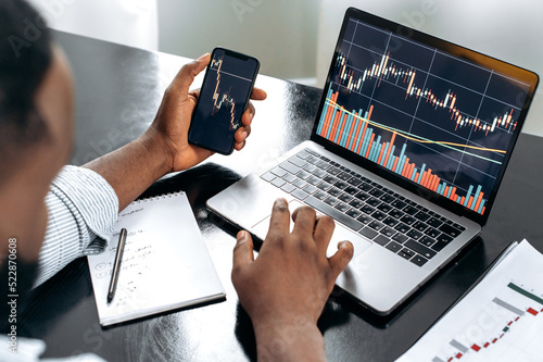 Crypto trader investor broker using cellphone and laptop for cryptocurrency financial market analysis, buying or selling cryptocurrency. Close-up of a laptop and cellphone screen with stock diagrams