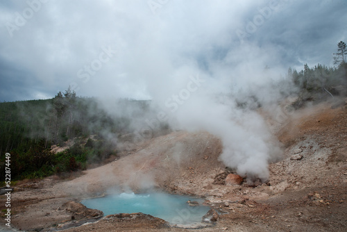 Active geyser in red dirt in a forest on a cloudy day in Yellowstone National Park Wyoming