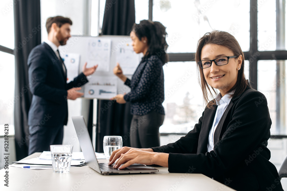 Confident elegant caucasian business woman with glasses, sitting at the desk at conference, near laptop. Beautiful elegant female employee on briefing meeting, looking at the camera, smiling friendly Confident elegant caucasian business woman with glasses, sitting at the desk at conference, near laptop. Beautiful elegant female employee on briefing meeting, looking at the camera, smiling friendly