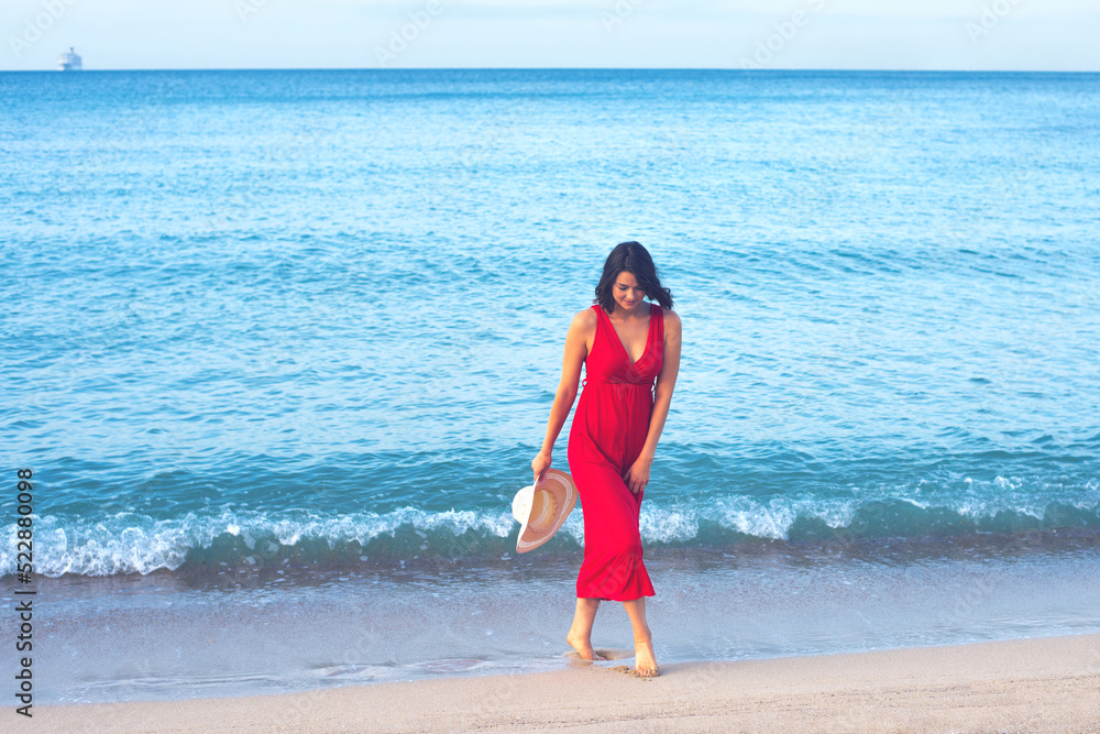 Young woman in red dress and white hat walking along the sand beach on a summer vacation 