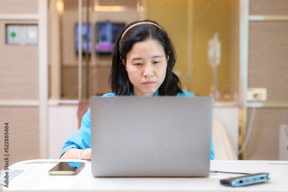 Asia patient woman sitting on the bed with saline solution in hospital ward using laptop, computer and check email. Working in clinic. Busy life of employee during sick. Healthcare and medical concept Asia patient woman sitting on the bed with saline solution in hospital ward using laptop, computer and check email. Working in clinic. Busy life of employee during sick. Healthcare and medical concept