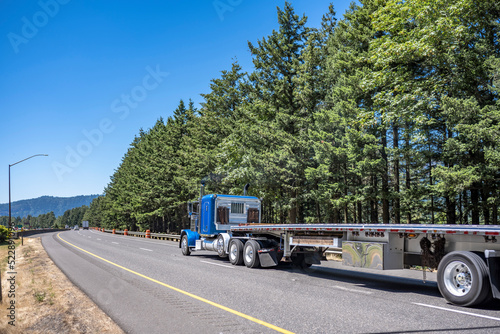 Classic blue big rig semi truck tractor with protect aluminum back wall and empty flat bed semi trailer moving on the wide highway interstate road
