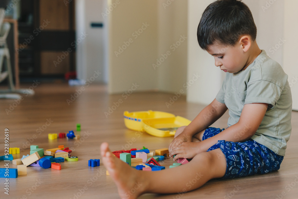Concentrated boy playing toys sitting on warm floor in modern living room. Baby development. Small house. Concentrated boy playing toys sitting on warm floor in modern living room. Baby development. Small house.