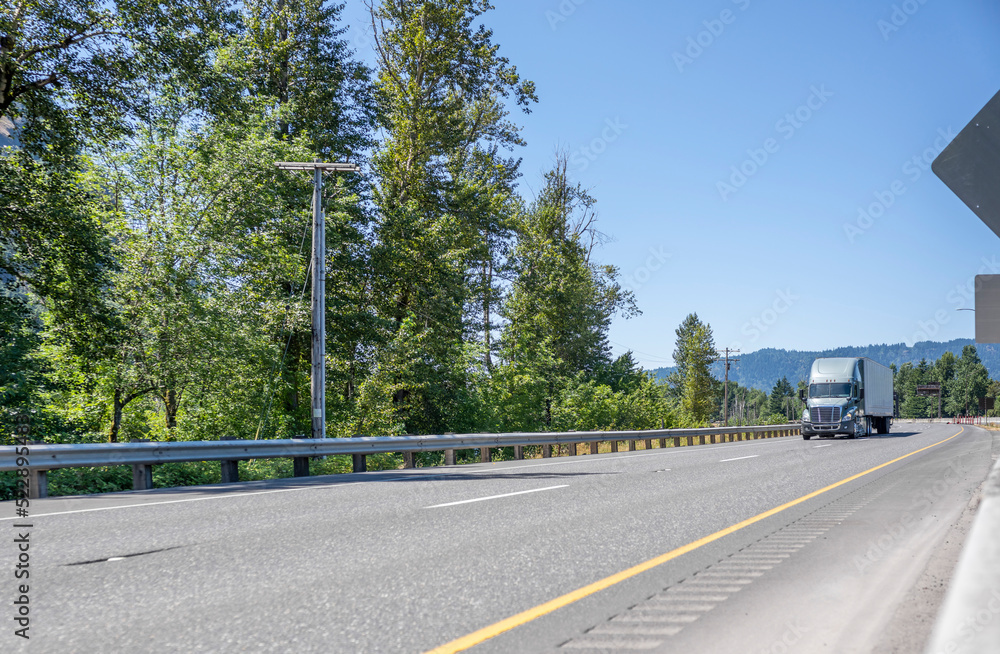 Light gray industrial grade big rig semi truck transporting cargo in dry van semi trailer moving on the wide highway road with green trees on the side