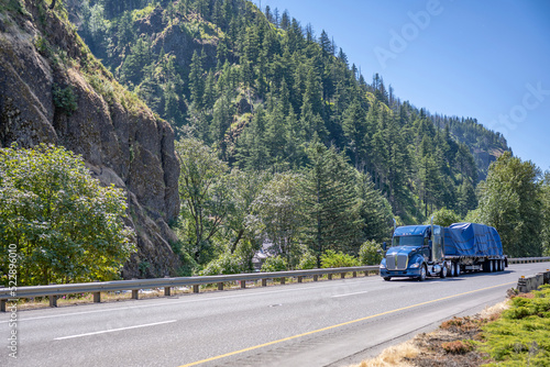 Powerful blue big rig semi truck transporting heavy duty cargo covered with tarp on flat bed semi trailer driving on the highway road in Columbia Gorge photo