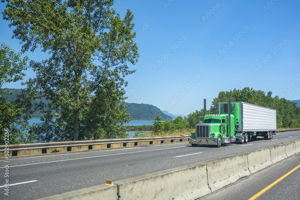 Stylish classic green big rig American semi truck with chrome accessories transporting cargo in the same style refrigerated semi trailer driving on the highway road along the Columbia River