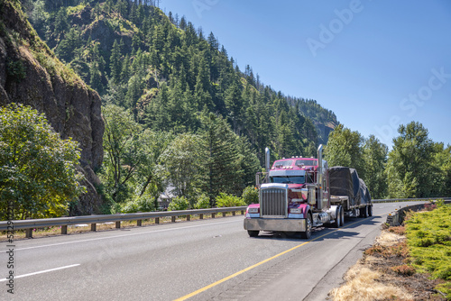 Stylish classic burgundy big rig American semi truck tractor transporting covered large cargo on step down semi trailer running on the highway road in Columbia Gorge