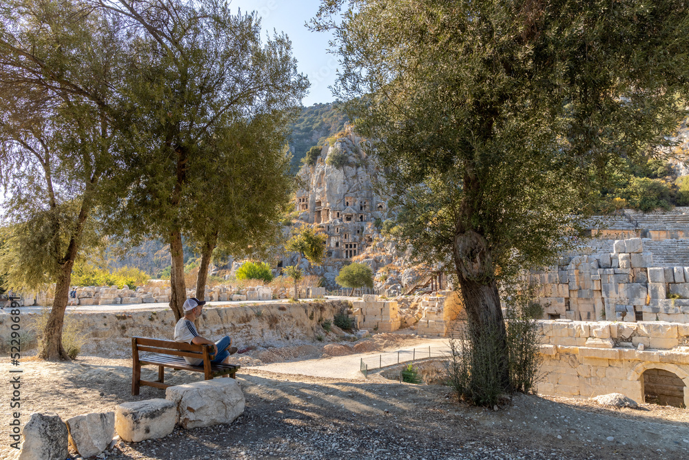Historical Myra ancient city. The man is sitting in the shade under the trees on a sunny summer day. Rock-cut tombs Ruins in Lycia region, Demre, Antalya, Turkey.  
