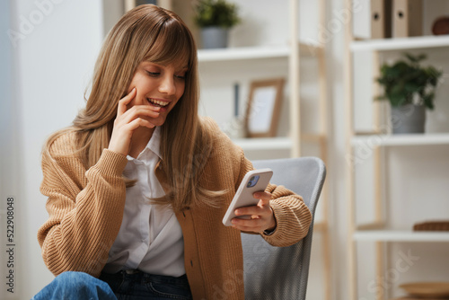 Cheerful young blonde woman in warm sweater get good news on studies by phone touch cheek sitting in armchair at home. Pause from work, take a break, social media in free time concept. Copy space