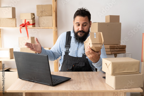 A male delivery service worker sits in overalls at the table and makes a video call. He spreads his arms to the sides, holds a crumpled box of goods. Delivery damage