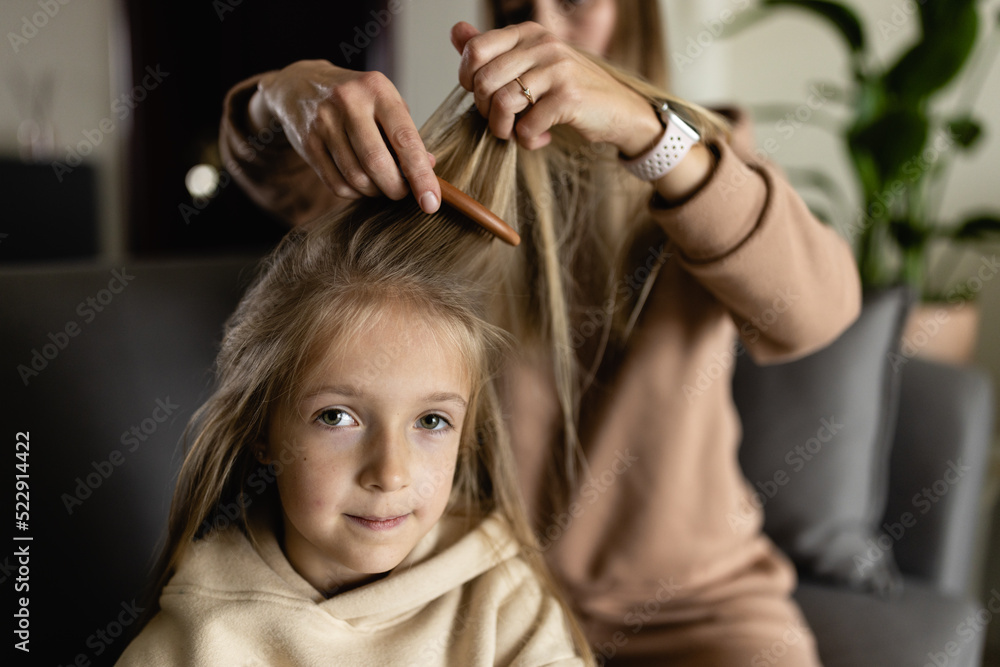 Fototapeta premium Charming adorable attractive beautiful caucasian young mother with blonde hair combing her small little schoolgirl daughter's hair at home