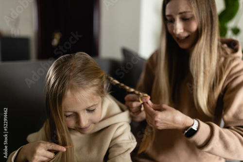 Charming adorable attractive beautiful caucasian young mother with blonde hair combing her small little schoolgirl daughter s hair at home