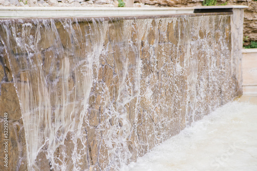 Artificial waterfall. Clear clear water falls against a background of gray stone or rock. Decoration of a park or garden.