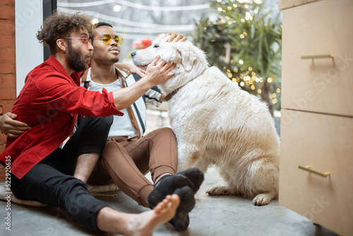 Two brightly dressed guys care dog while sitting on the floor together indoors on background of backyard. Concept of gay couples and romance. Caucasian and hispanic man with pet at home
