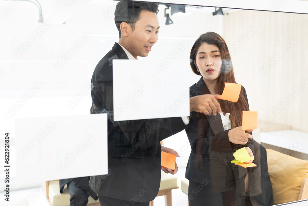 businesspeople office worker reading sticky notes on planning glass board. Business team pointing on keywords in sticky notes and business report to brainstorming about planning strategy of business. businesspeople office worker reading sticky notes on planning glass board. Business team pointing on keywords in sticky notes and business report to brainstorming about planning strategy of business.