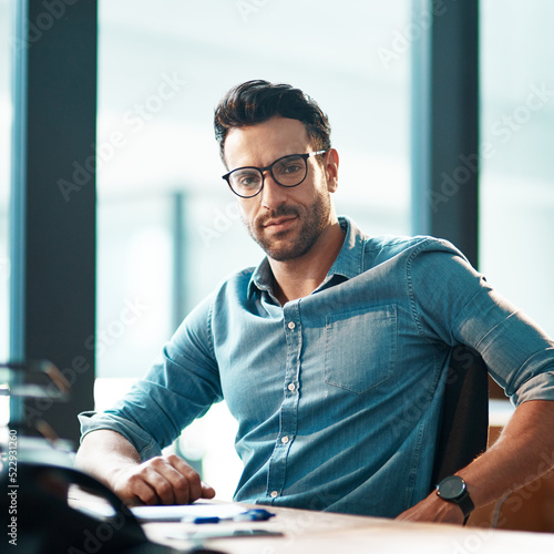 Billede på lærred Young, confident and professional business man wearing glasses, sitting at his desk and working in a modern office portrait