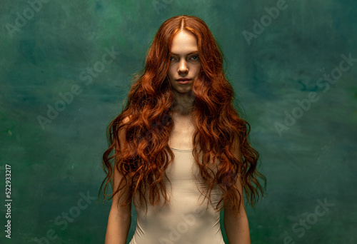 Adorable tender redhead girl with long curly hair isolated over dark green background. Fabolous curly hair and harming gaze. Concept of beauty, art photo