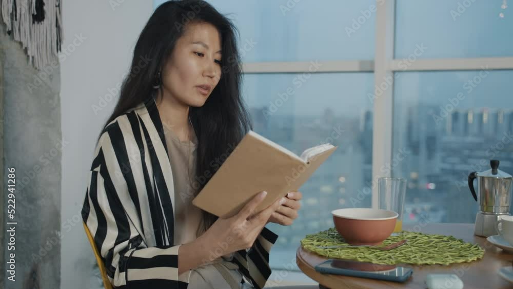 Beautiful Asian woman reading book and drinking orange juice enjoying evening in apartment alone. Healthy lifestyle and leisure time concept. Beautiful Asian woman reading book and drinking orange juice enjoying evening in apartment alone. Healthy lifestyle and leisure time concept.