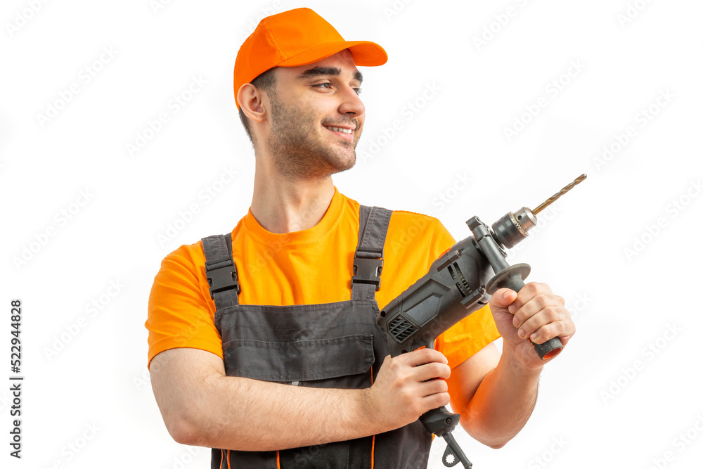 Young worker drilling with electric drill in his hands. Tool for boring holes. Craftsman repairman wearing orange uniform cap and t-shirt, overalls on a white isolated background for advertising Young worker drilling with electric drill in his hands. Tool for boring holes. Craftsman repairman wearing orange uniform cap and t-shirt, overalls on a white isolated background for advertising