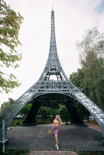 Fototapeta Naklejka Na Ścianę i Meble -  A stylish slender smiling girl with blond long hair stands with a bouquet of wild flowers in her hands, near a copy of the Eiffel Tower in Ukraine. Dressed in a bright summer skirt and a sexy top.