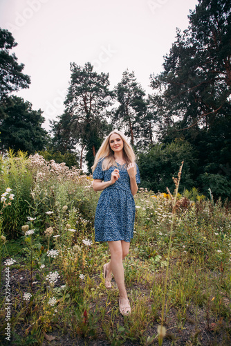 A portrait of a beautiful slender girl with blond long hair holding wildflowers in her hands, in a blue summer dress with a floral pattern, walks along a blooming meadow in summer.