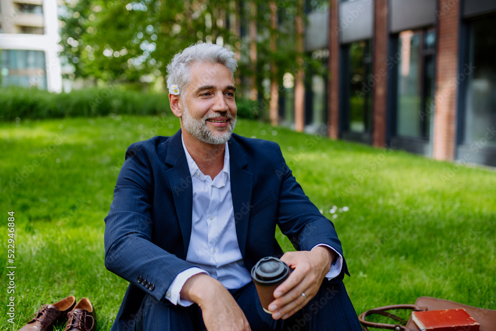 Obraz premium Mature businessman resting and sitting barefoot in park with cup of coffee, feeling free, escaping from work, work life balance concept.