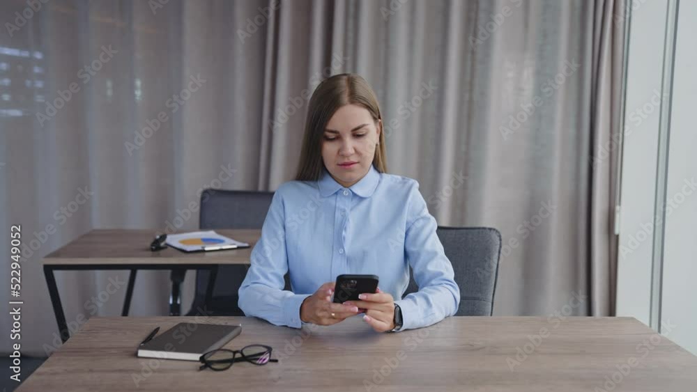 Long-haired lady wearing blue shirt sits at desk. Woman looks at her phone and takes notes into her paper notebook. Long-haired lady wearing blue shirt sits at desk. Woman looks at her phone and takes notes into her paper notebook.