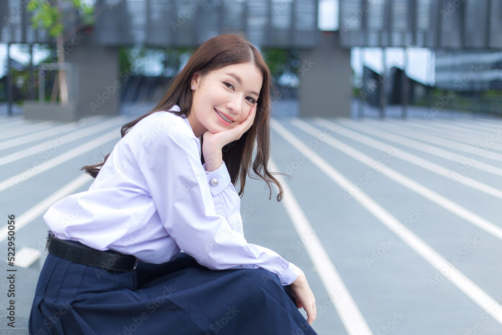 Beautiful Asian high school student girl in the school uniform with braces on her teeth sits and smiles confidently while she looks at the camera happily with the building in the background. Beautiful Asian high school student girl in the school uniform with braces on her teeth sits and smiles confidently while she looks at the camera happily with the building in the background.