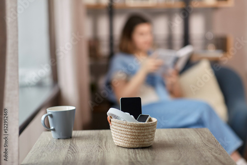 digital detox and leisure concept - close up of gadgets in basket on table and woman reading book at home